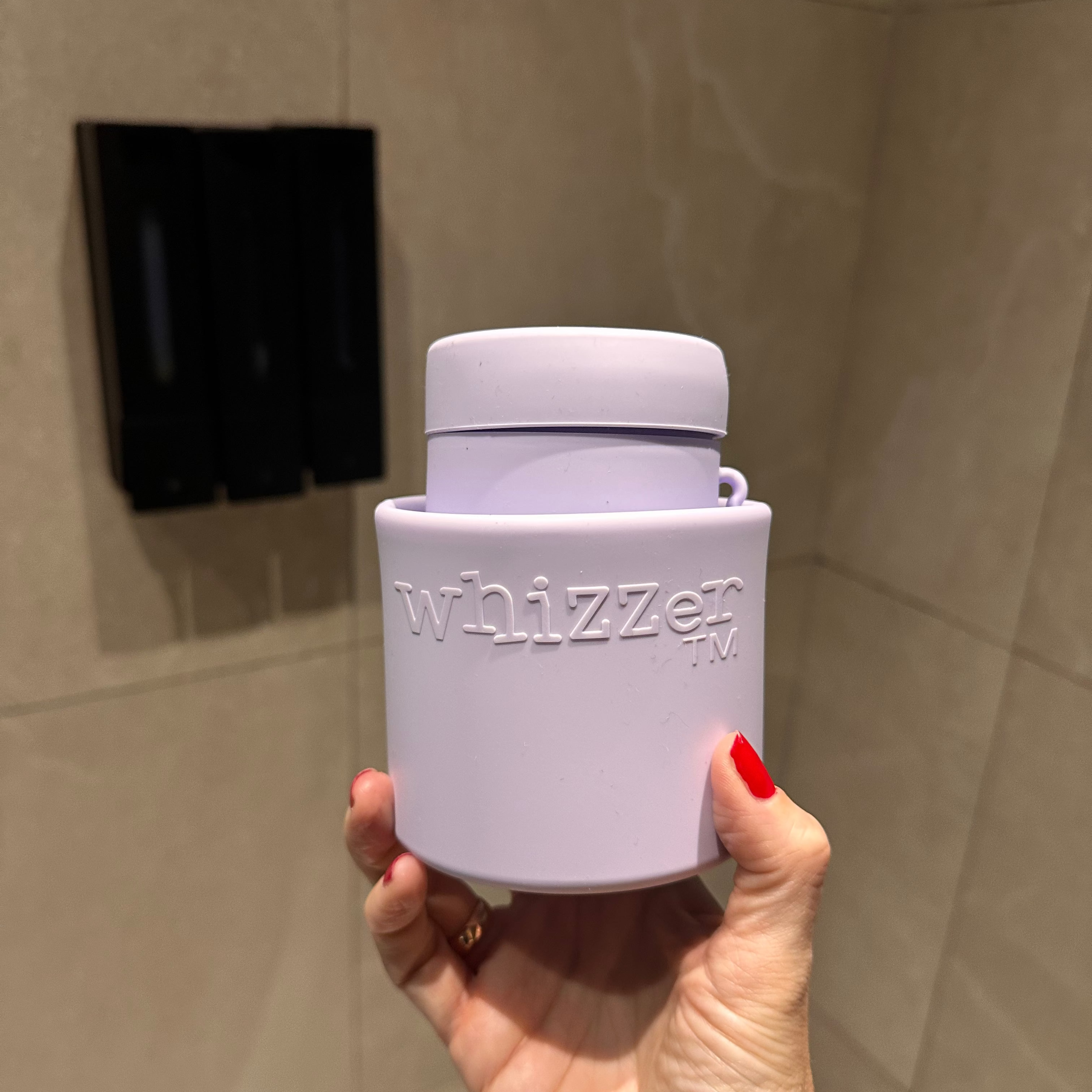 Hand holding a purple 'whizzer' container against a tiled bathroom wall.
